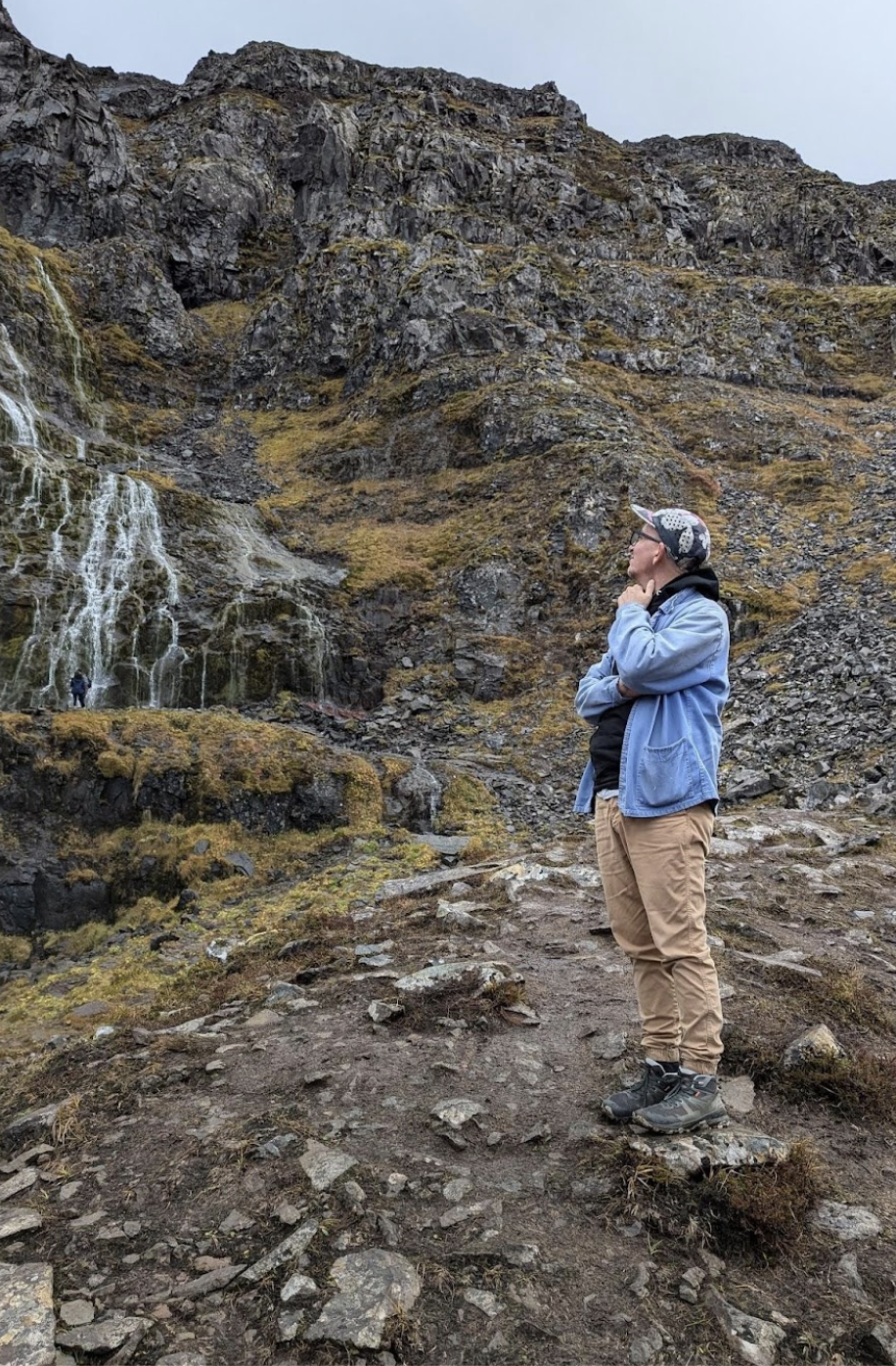 A man in tan pants and a light blue jacket standing in front of a waterfall in Iceland